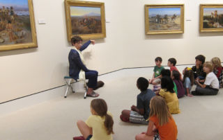 woman talking in front of a painting to a group of children seated on floor of museum gallery
