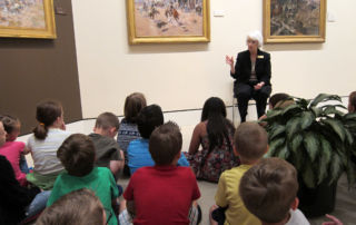 woman talking about painting and seated in front of group of children in museum gallery