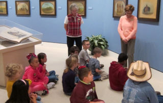 two adults discussing artworks with a group of children seated on the floor