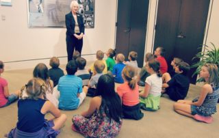 white woman standing in front of and talking to a group of children seated on floor