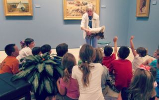 white woman showing model covered wagon to group of students in museum gallery