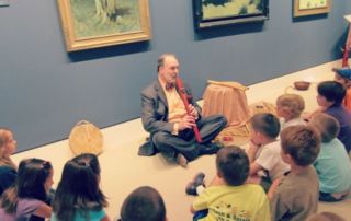 man sitting crossed-legged on floor playing wooden flute to group of young kids