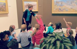 woman pointing to boy holding up hand among a group of kids in a museum gallery