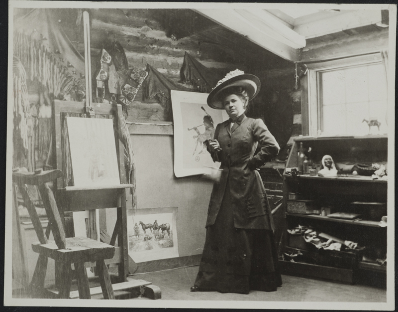 a woman in a dress and big hat standing in an artist studio