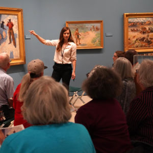 a woman standing in front of a group of people pointing to a painting