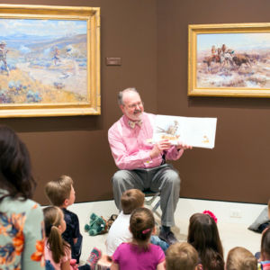 man in pink shirt in front of paintings reading a book to a group of kids