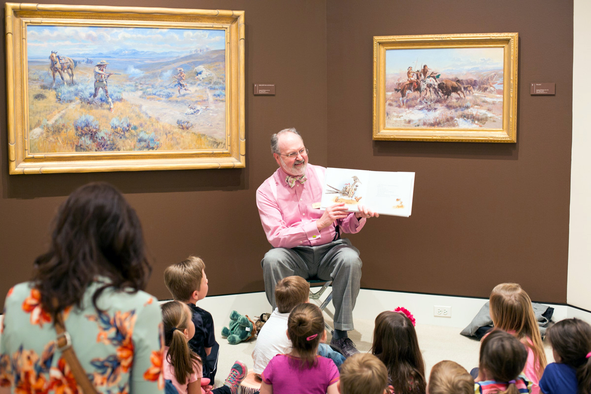 man in pink shirt in front of paintings reading a book to a group of kids