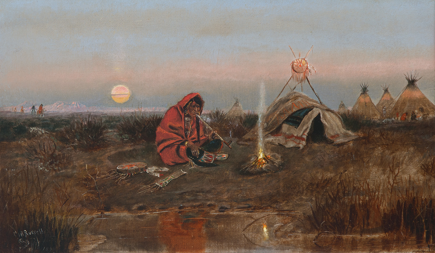 A Cree man sits by a fire smoking a pipe with tents in the distance.