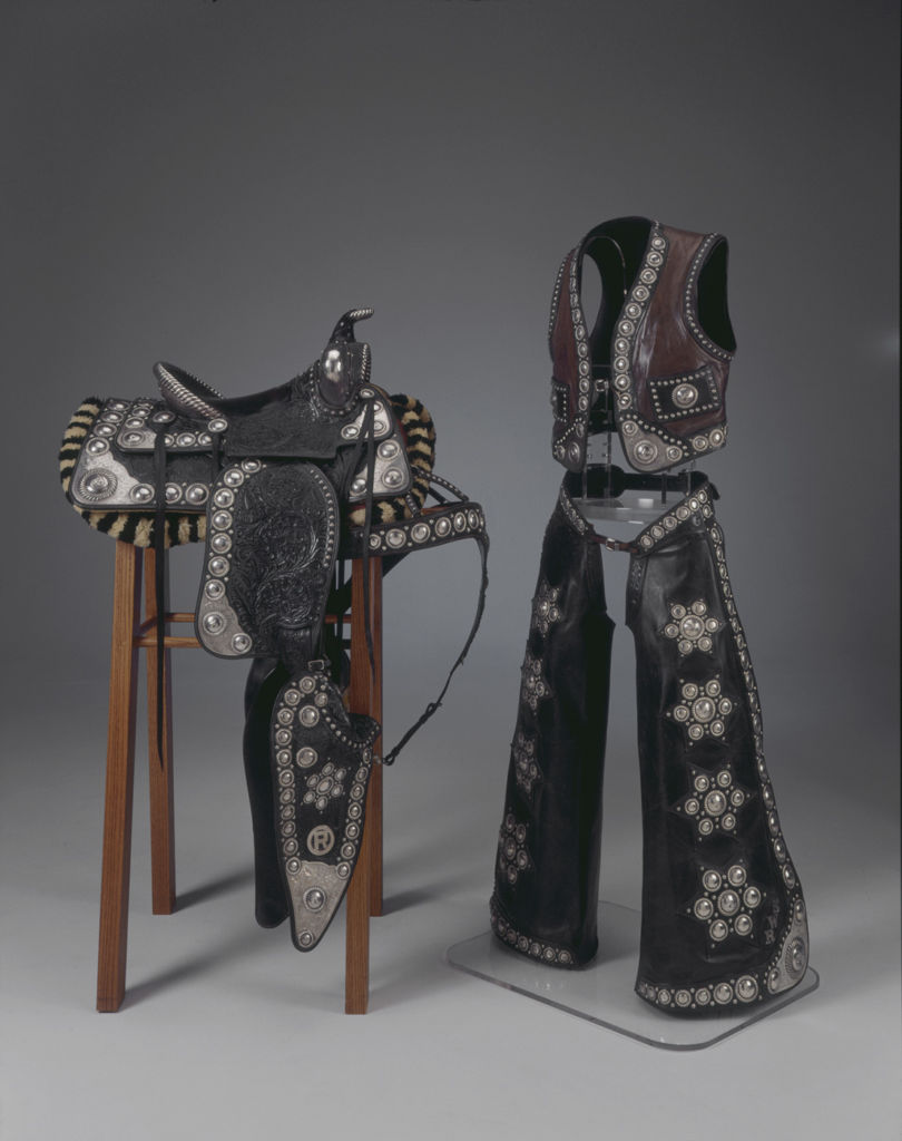 A black leather saddle decorated with sterling silver on a wooden armature next to a matching vest and chaps.