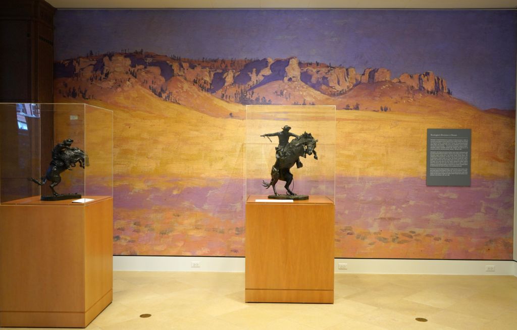 a couple of bronze sculptures inside vitrines placed in front of a photo mural of a painted landscape