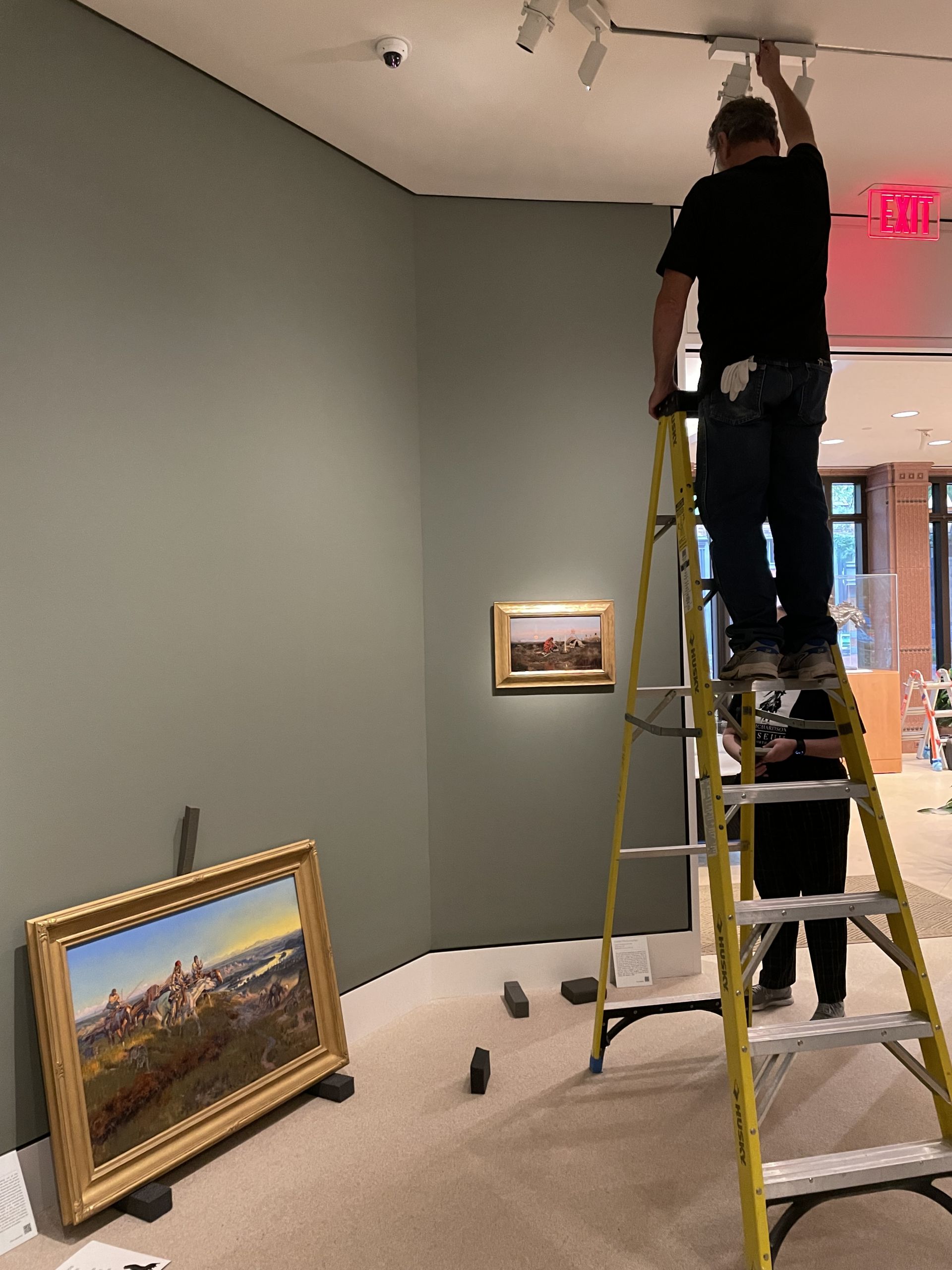 A man on a ladder adjusting a light fixture while looking at a painting on the wall below.