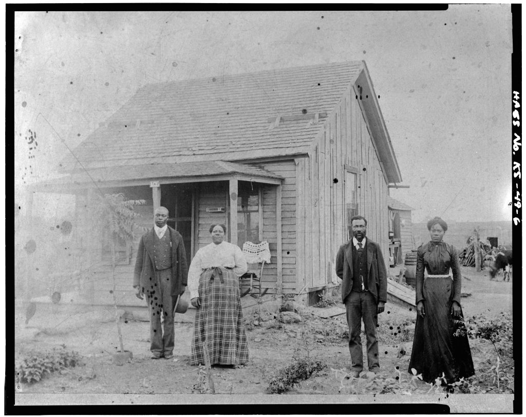 A Black family posing in front of a small, wooden home on the plains.