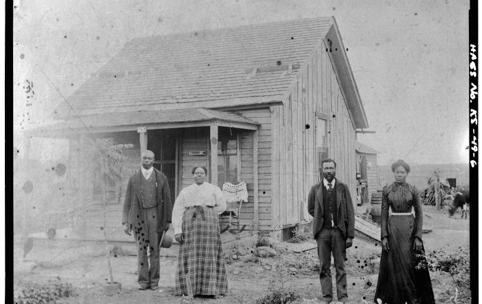 A Black family posing in front of a small, wooden home on the plains.