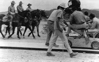 film director behind camera recording three men on horseback moving toward the camera
