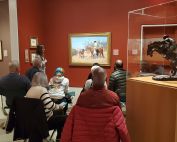 A group of people sitting in front of and talking about a painting in a museum gallery.
